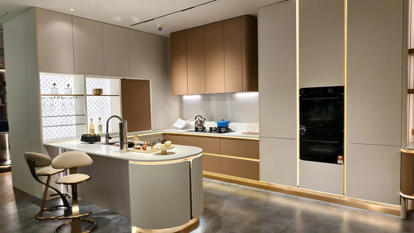 Pale grey and warm taupe handleless kitchen with a curved island, integrated oven bank and under-cabinet shadow line