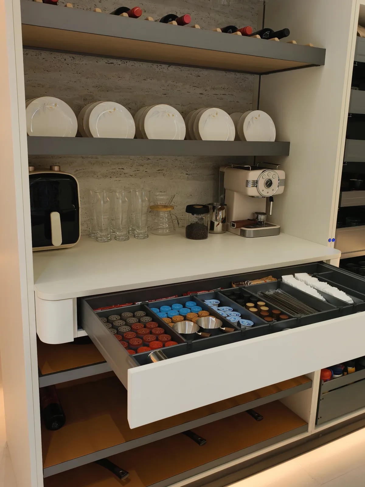 Tall white kitchen unit with a drawer pulled out showing compartments of red, blue and brown coffee capsules, sugar sachets and stirrers