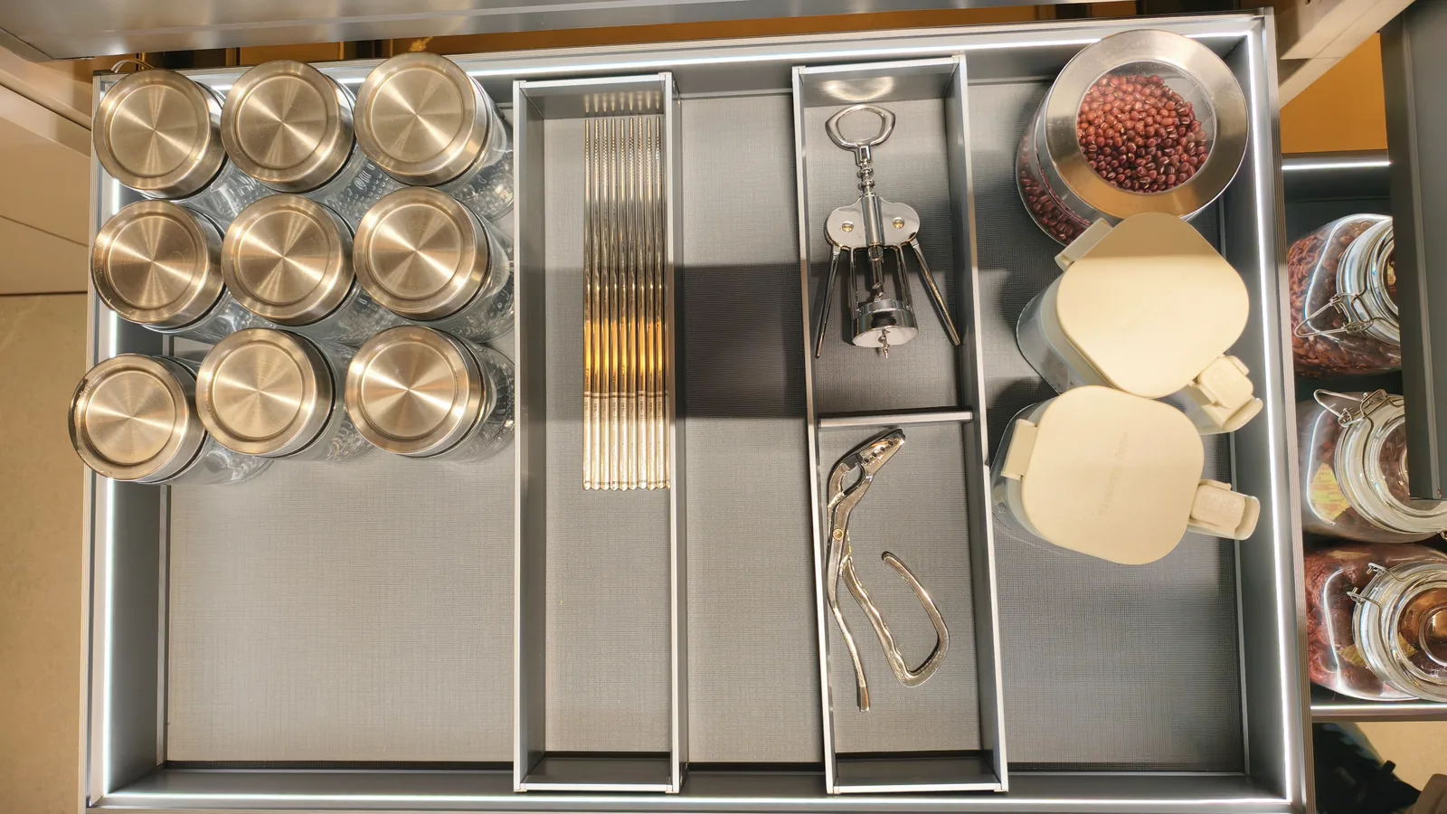 Open kitchen drawer with factory-cut compartments holding glass spice jars, chopsticks, a corkscrew and a jar of red beans