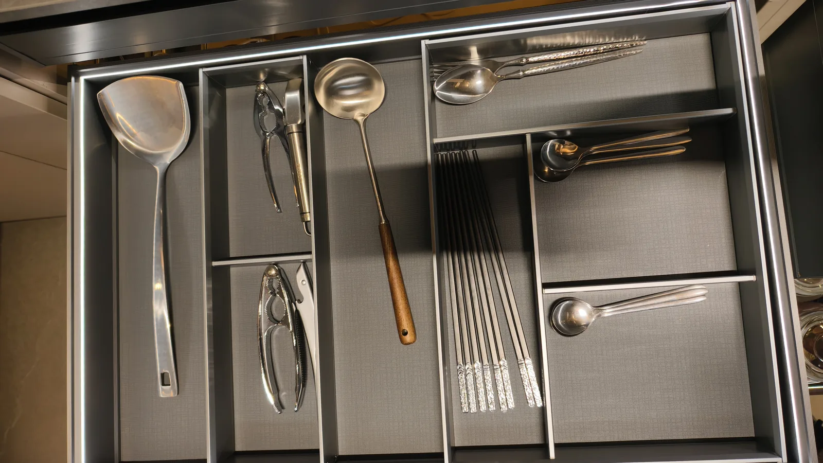 Top-down view of a kitchen drawer divided into factory-cut compartments holding a wok spatula, nutcracker, ladle, chopsticks and spoons
