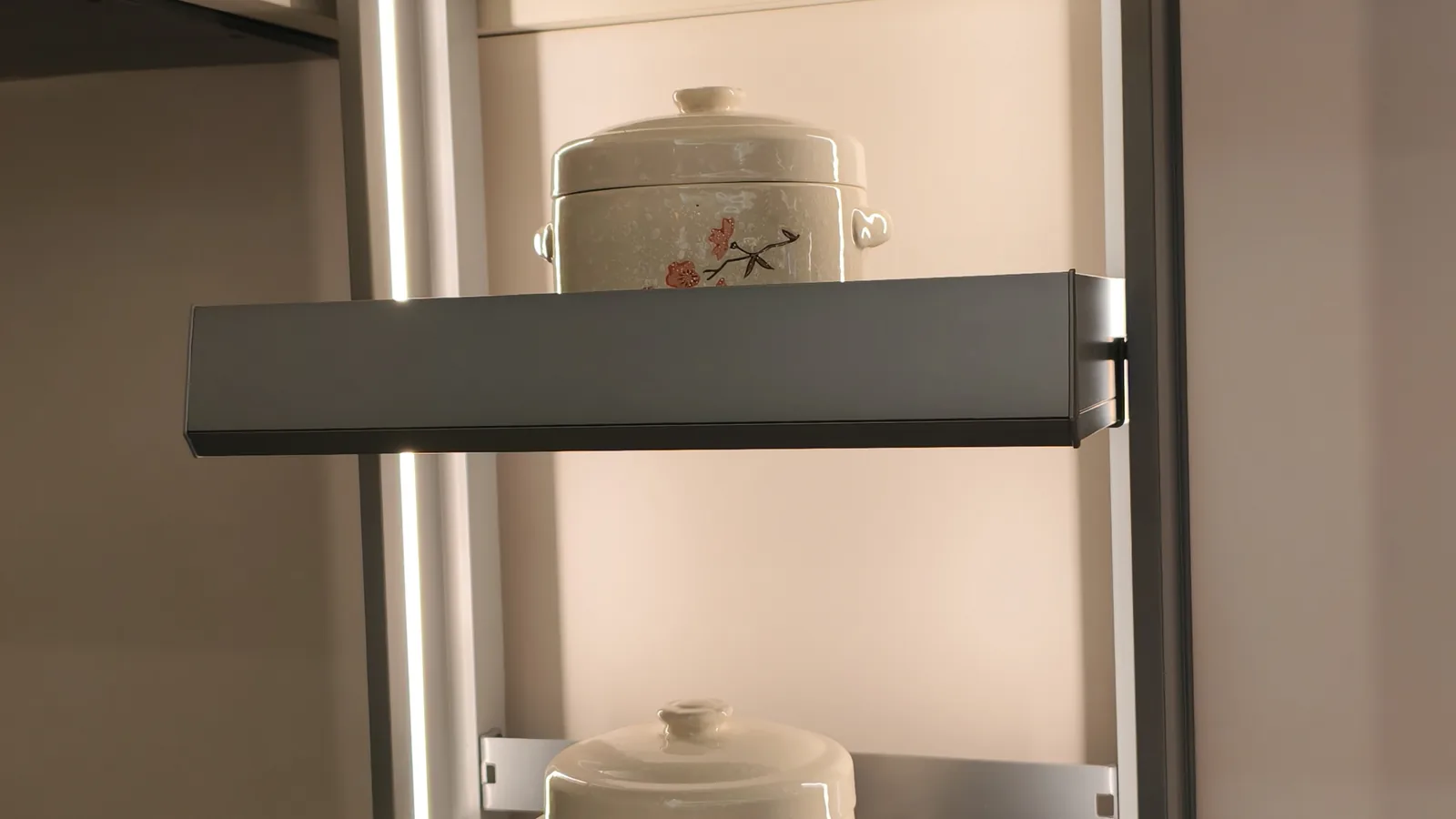 Pull-down basket shelf lowered out of a wall cabinet, loaded with a covered ceramic stew pot, lit by an LED strip inside the cabinet